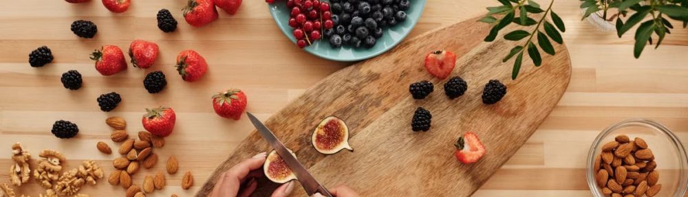 Overhead shot of fresh fruits, nuts, and hands preparing ingredients on a wooden board.